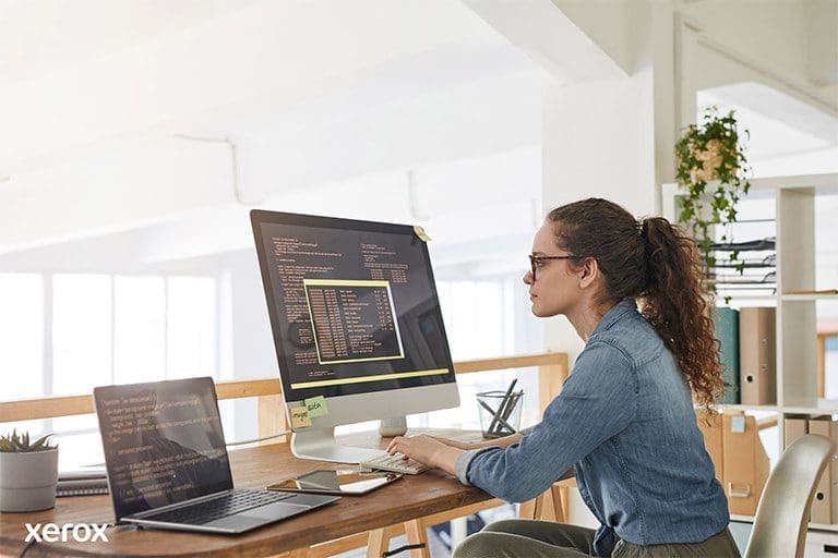 Eine Frau an ihrem Schreibtisch, beschäftigt mit einem Computer und einem Laptop, demonstriert Produktivität an einem modernen Arbeitsplatz. Eine Frau an ihrem Schreibtisch, beschäftigt mit einem Computer und einem Laptop, demonstriert Produktivität an einem modernen Arbeitsplatz.