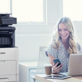 Eine Person schaut zufrieden auf ihr Smartphone, w&auml;hrend sie mit einem Laptop an einem Schreibtisch sitzt. Neben dem Schreibtisch in einem hell erleuchteten B&uuml;roraum ist ein Multifunktionsdrucker Xerox VersaLink B605 zu sehen.