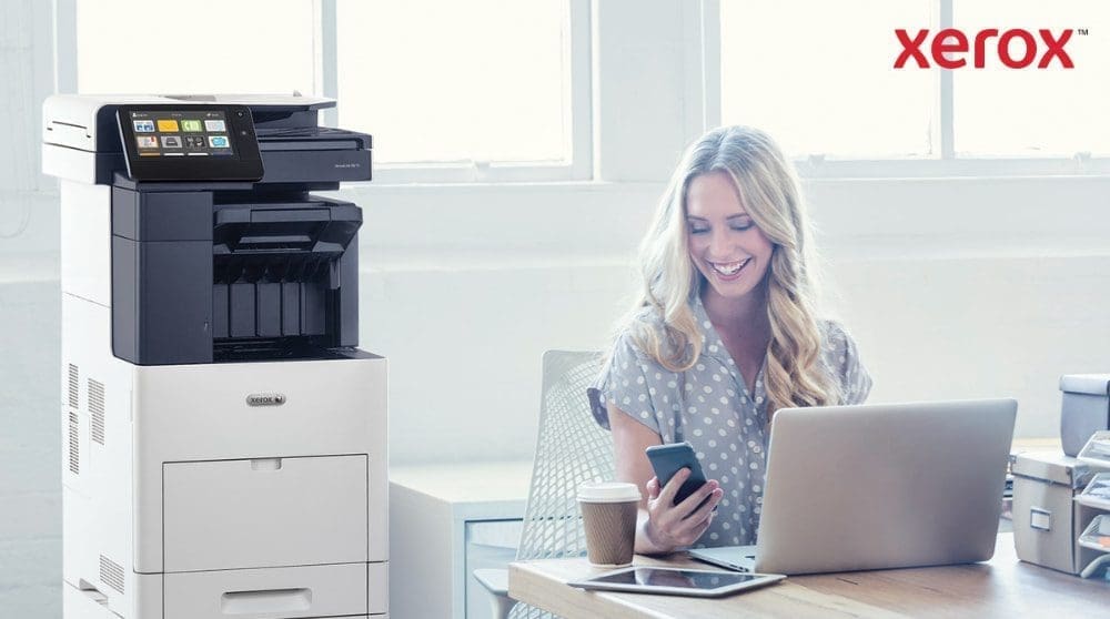 Eine Person schaut zufrieden auf ihr Smartphone, w&auml;hrend sie mit einem Laptop an einem Schreibtisch sitzt. Neben dem Schreibtisch in einem hell erleuchteten B&uuml;roraum ist ein Multifunktionsdrucker Xerox VersaLink B605 zu sehen.