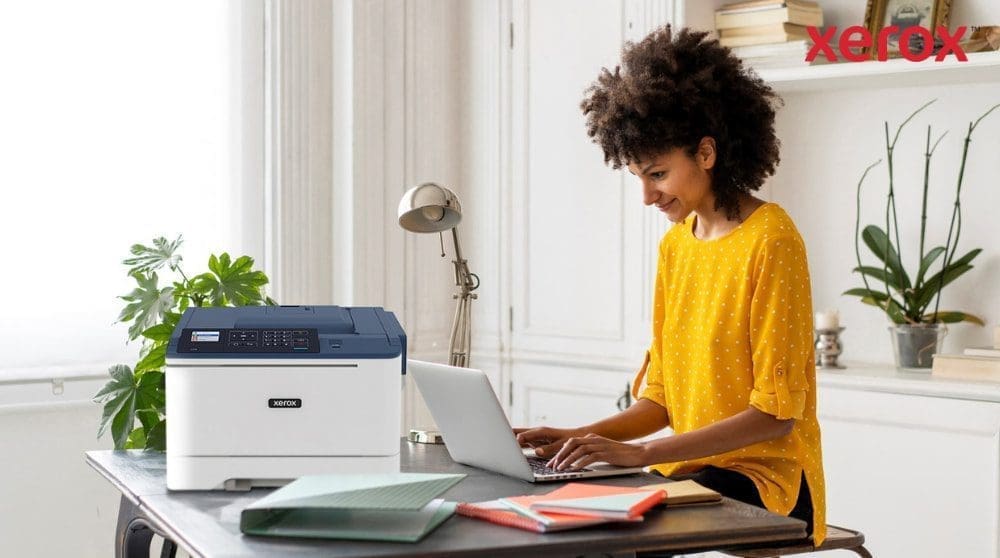 Eine Person mit lockigem Haar lächelt, während sie an einem Schreibtisch auf einem Laptop tippt. Neben dem Laptop steht ein kompakter Xerox C310-Drucker, der ein modernes Home-Office-Setup für mehr Produktivität vorführt.