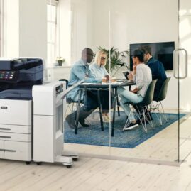 Im Vordergrund eines hellen B&uuml;ros steht ein moderner Multifunktionsdrucker Xerox AltaLink B8245. Im Hintergrund sitzt eine Gruppe von Menschen an einem Tisch und diskutiert gemeinsam.
