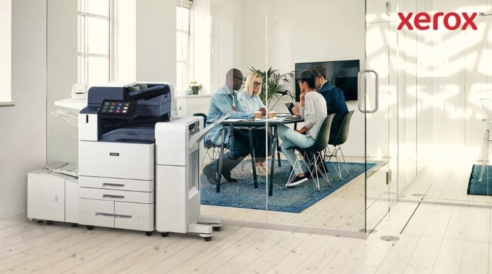 Im Vordergrund eines hellen B&uuml;ros steht ein moderner Multifunktionsdrucker Xerox AltaLink B8245. Im Hintergrund sitzt eine Gruppe von Menschen an einem Tisch und diskutiert gemeinsam.