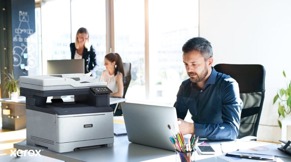 Xerox® Smart Start: Entwickelt für moderne Arbeitsplätze Eine Person sitzt an einem Schreibtisch und blickt auf einen Laptop, daneben steht ein Xerox C325 Multifunktionsdrucker. Im Hintergrund arbeiten zwei weitere Personen ebenfalls an ihren Schreibtischen in einer hellen Büroumgebung.