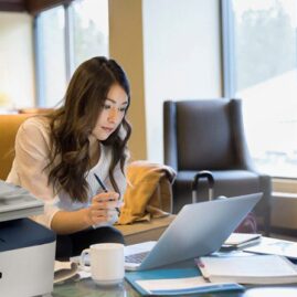 Eine Frau arbeitet in einem gem&uuml;tlichen B&uuml;ro oder einer Lounge an einem Laptop. Im Vordergrund ist ein kompakter Xerox-Drucker zu sehen, der auf eine nahtlose Integration des Druckens in digitale Arbeitsabl&auml;ufe hinweist.