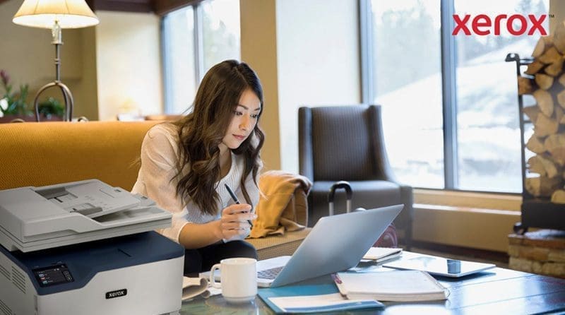 Eine Frau arbeitet in einem gem&uuml;tlichen B&uuml;ro oder einer Lounge an einem Laptop. Im Vordergrund ist ein kompakter Xerox-Drucker zu sehen, der auf eine nahtlose Integration des Druckens in digitale Arbeitsabl&auml;ufe hinweist.