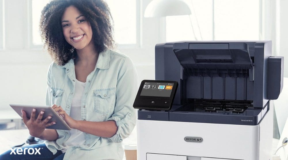Xerox® Workplace Cloud und Suite: Benutzerfreundliche Integration Eine lächelnde Person sitzt neben einem weißen Xerox VersaLink B610-Drucker, hält ein Tablet und blickt in die Kamera, was die Benutzerfreundlichkeit der Xerox-Lösungen hervorhebt.