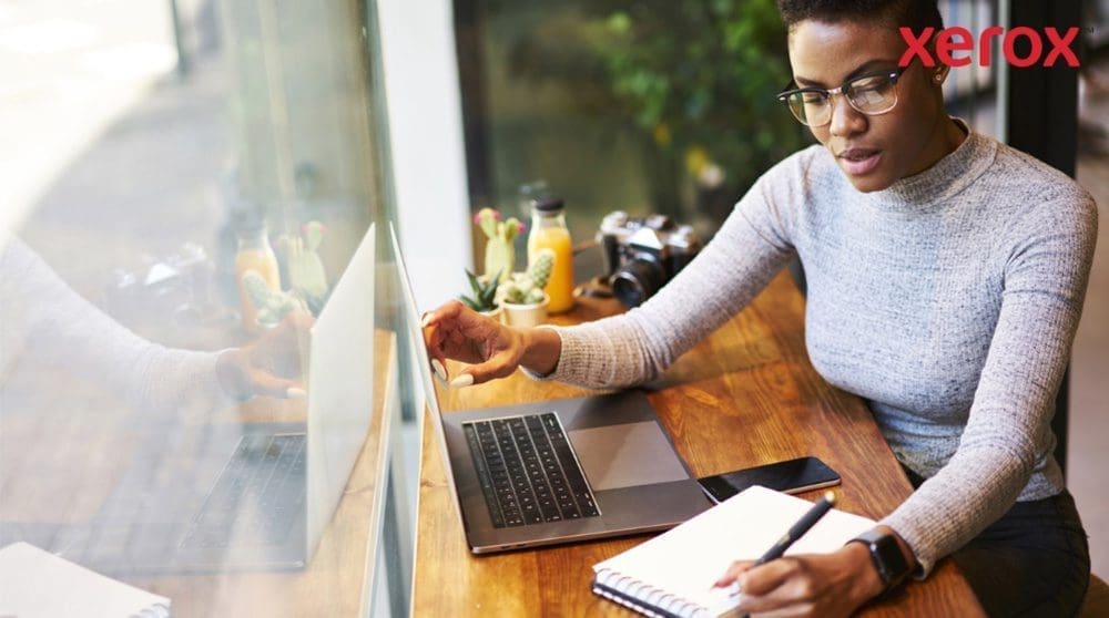 Xerox® Workplace Cloud und Suite: Verbesserte Produktivität Eine Person mit Brille arbeitet an einem Holztisch, schaut auf einen Laptop-Bildschirm und schreibt in ein Notizbuch, während ein Smartphone in der Nähe ist, was auf eine produktive Arbeitsumgebung hindeutet