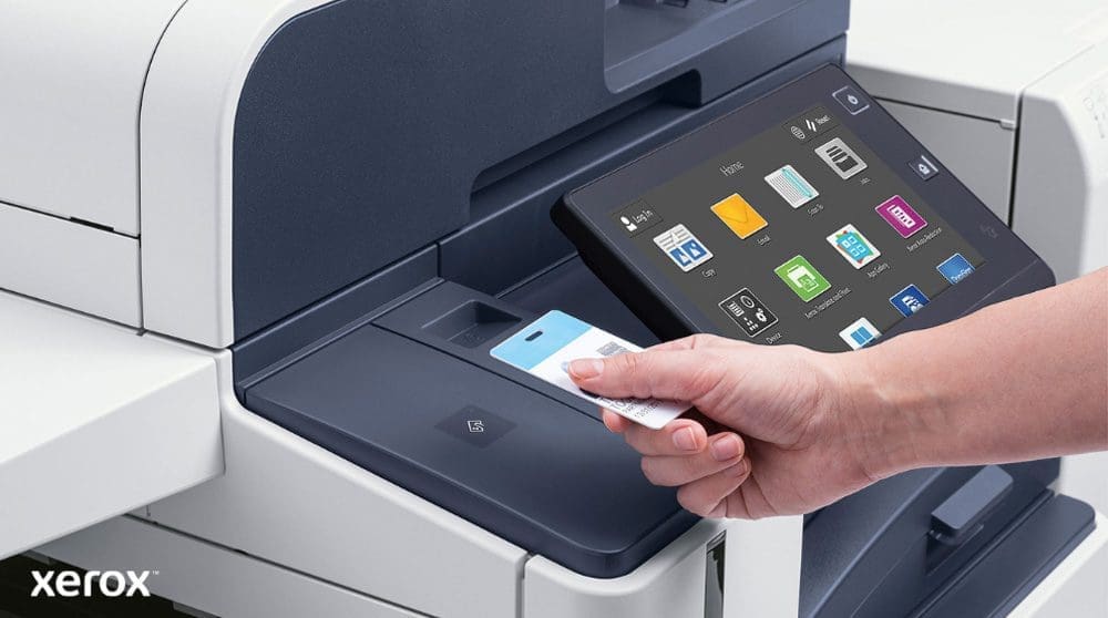 Xerox® Workplace Cloud und Suite: Sichere Authentifizierung und intelligente Schnittstelle Eine Hand hält eine Sicherheitskarte und scannt sie in der Nähe des Kartenlesers eines modernen Xerox-Multifunktionsdruckers mit einem großen Touchscreen-Display, auf dem verschiedene Anwendungssymbole angezeigt werden.
